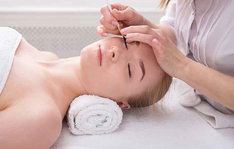 woman gets eyelashes tinting by beautician at spa